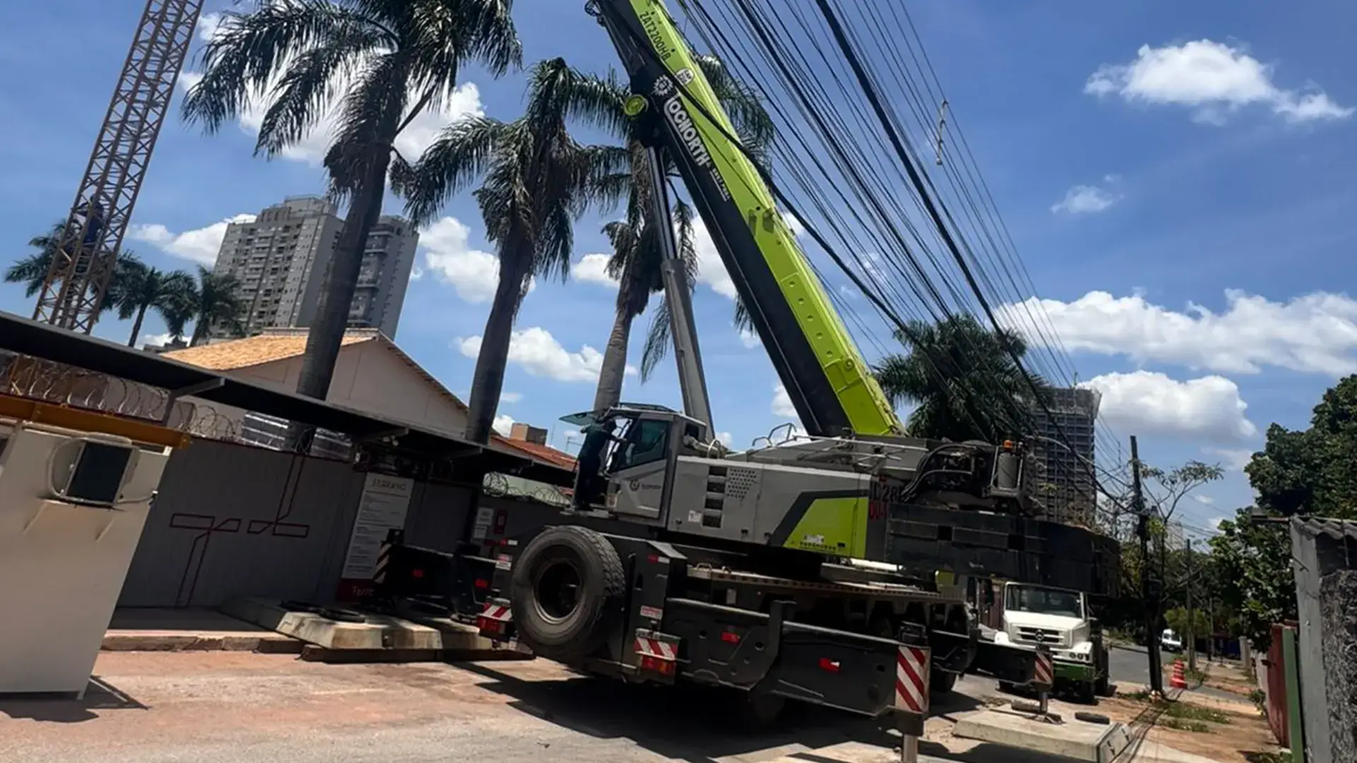 Guindaste móvel verde patolado em uma via urbana, operando com a lança estendida próxima a fios de energia elétrica e palmeiras.
