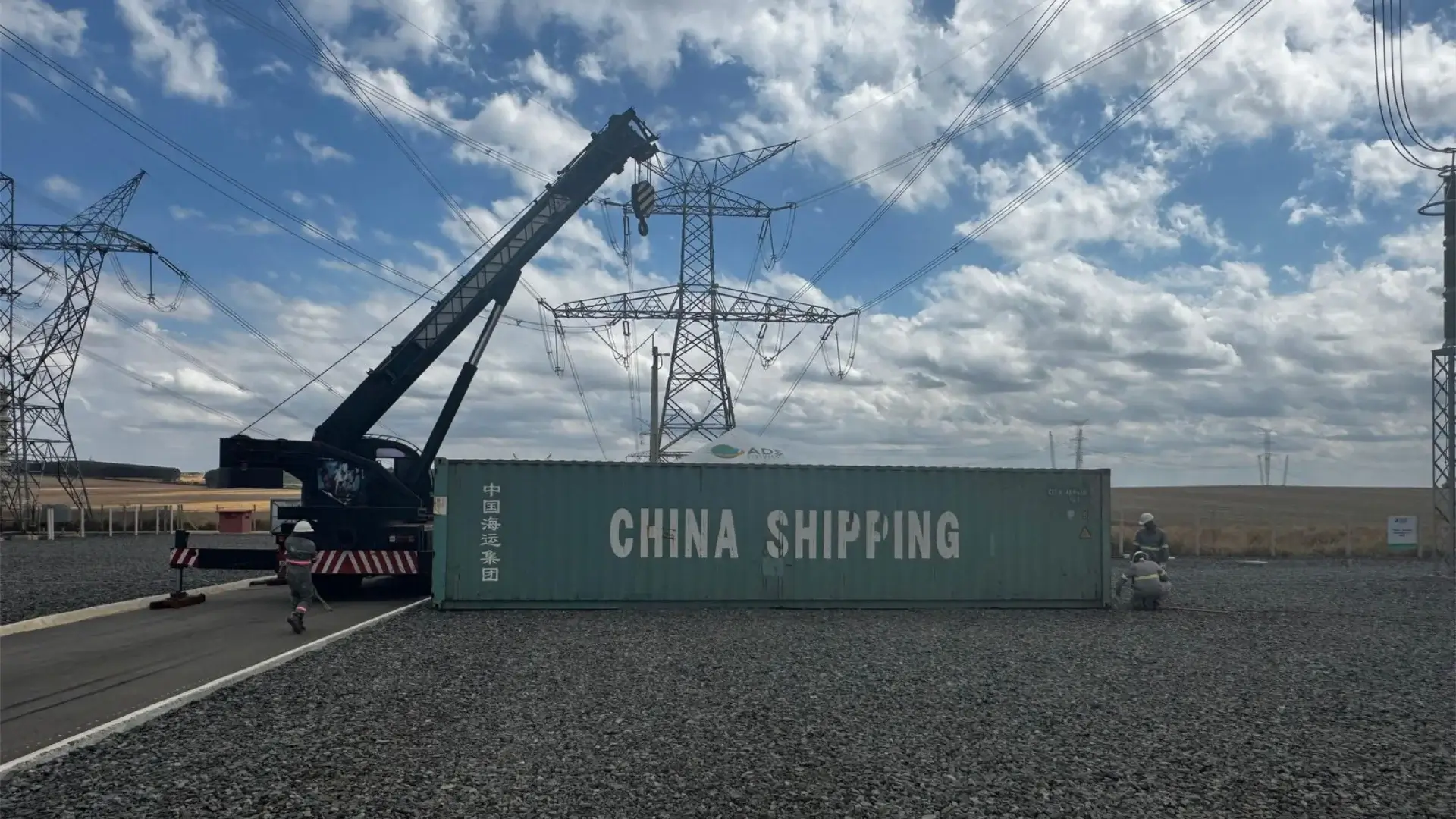 Um guindaste móvel preto posicionado para içar um contêiner de transporte verde-azulado da "China Shipping" em um pátio de infraestrutura com torres de transmissão de energia e trabalhadores.